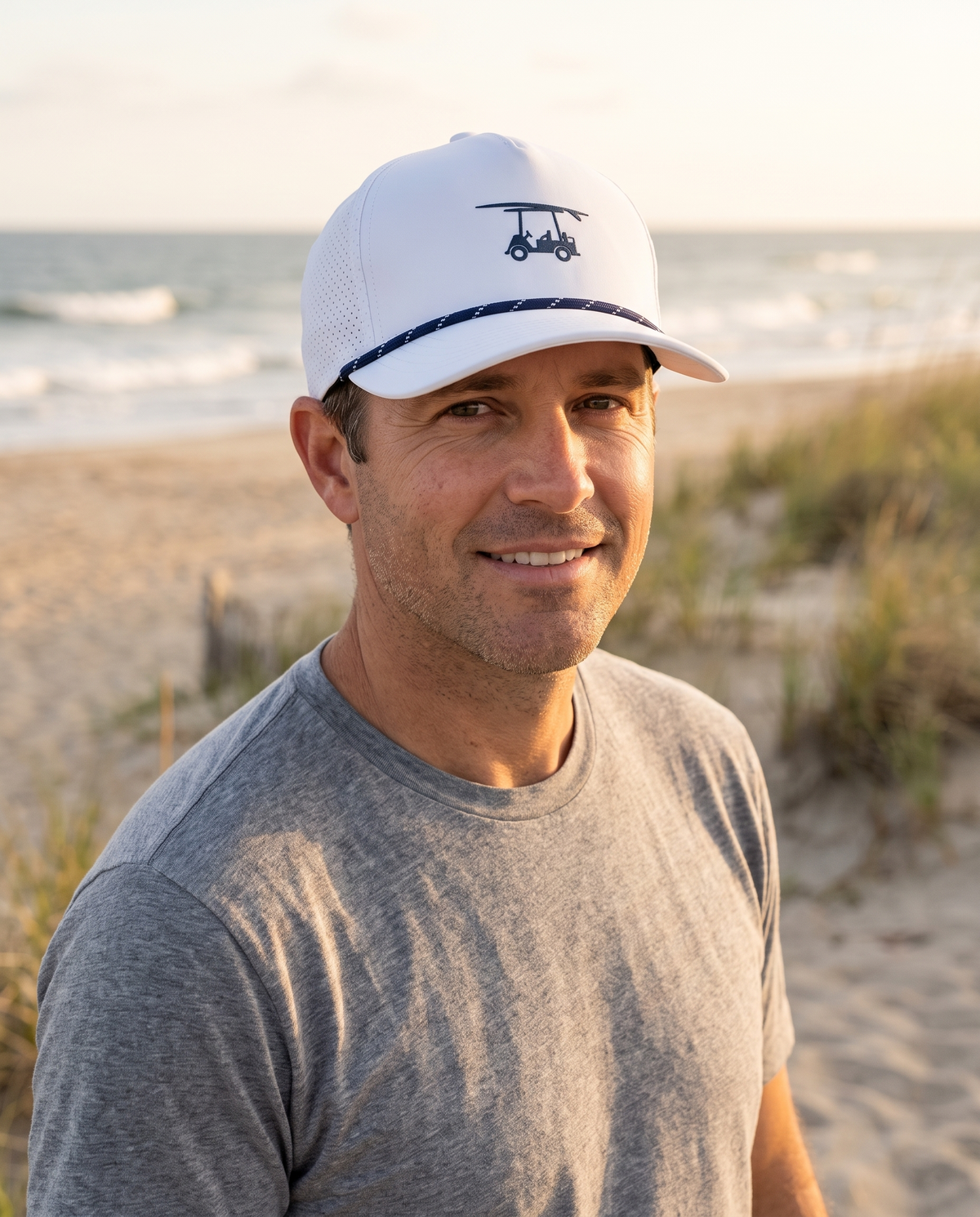 Man on the dunes above the surf wearing the Bald Head Blues Performance Rope Hat in White with the navy cart and surfboard mark
