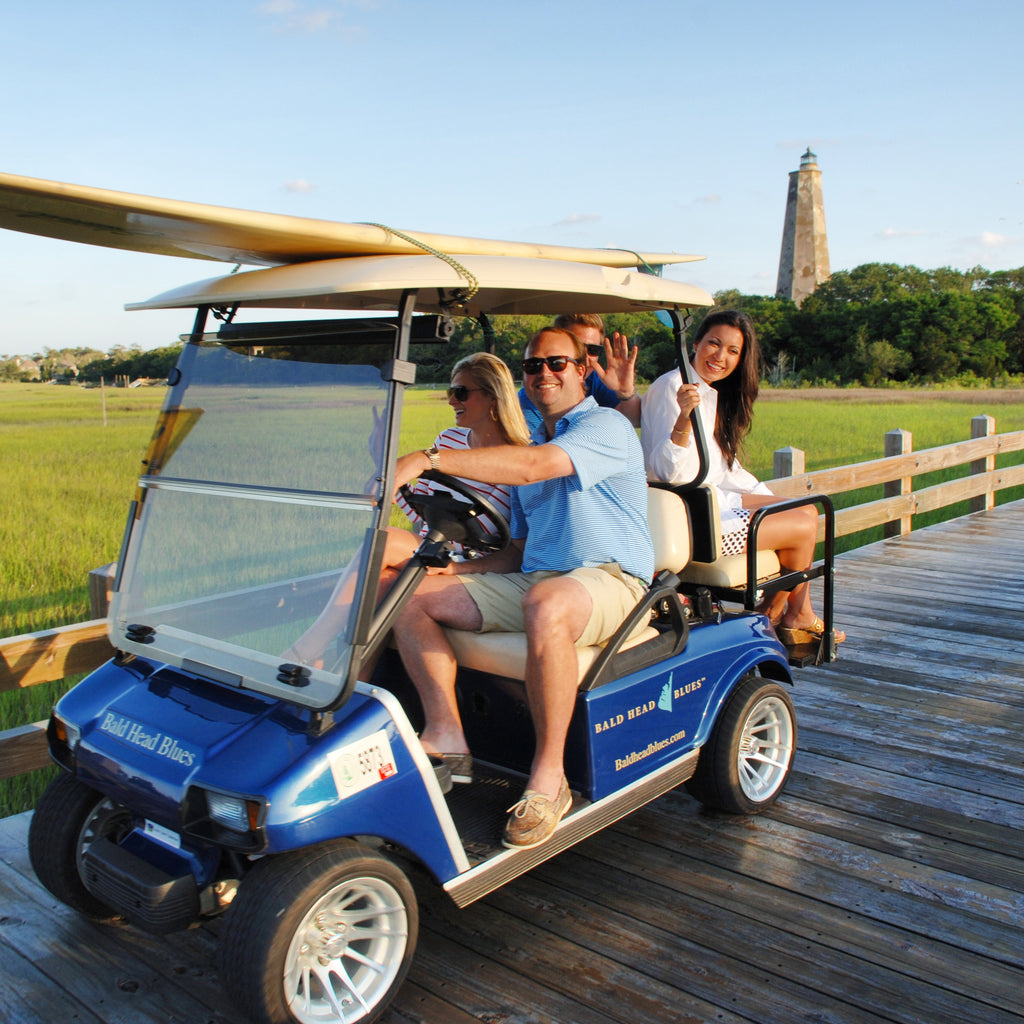 The family riding a Bald Head Blues golf cart with a surfboard on the roof, Old Baldy lighthouse behind