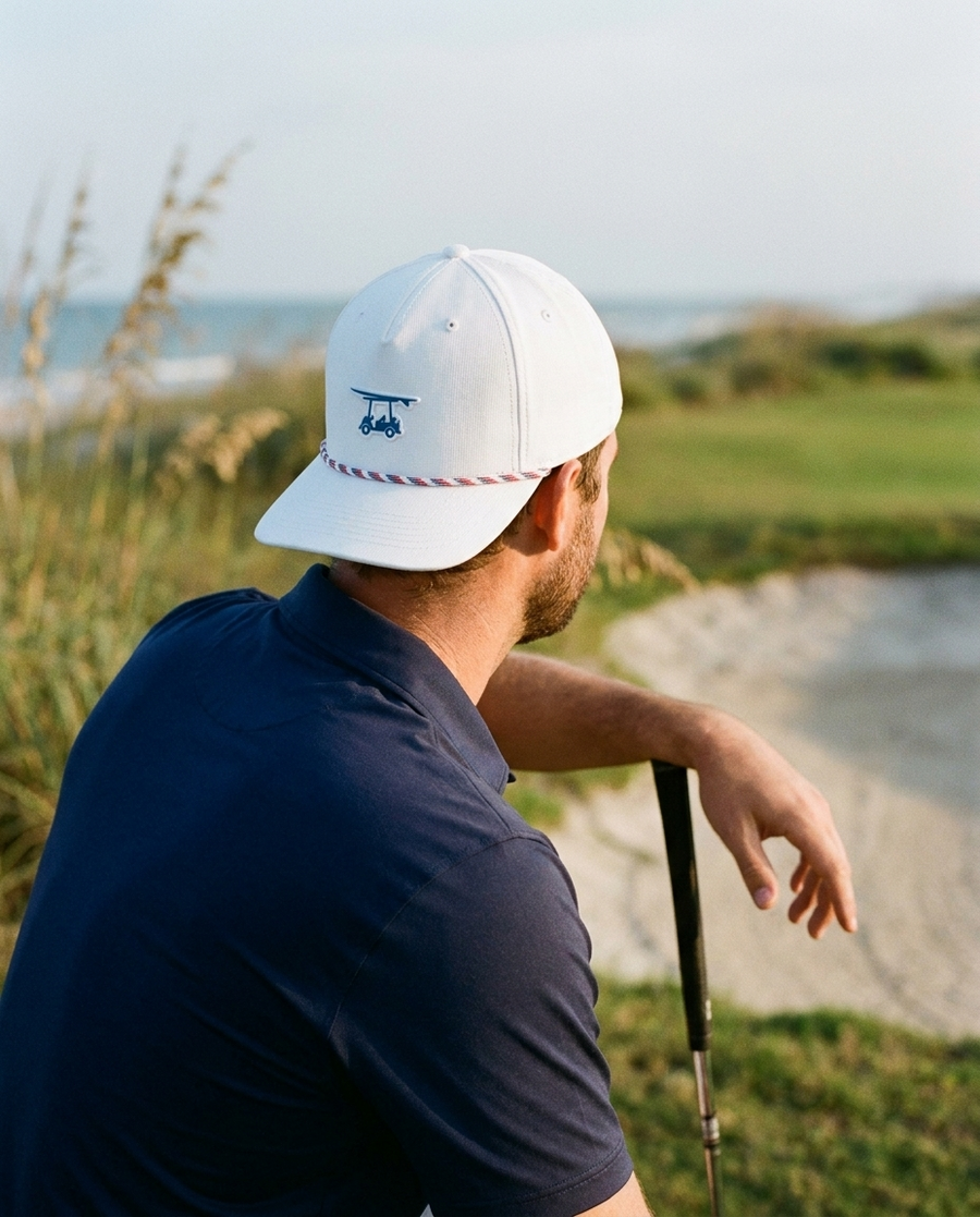 Golfer in a white five-panel roped cap leaning on a putter above a bunker on a coastal course