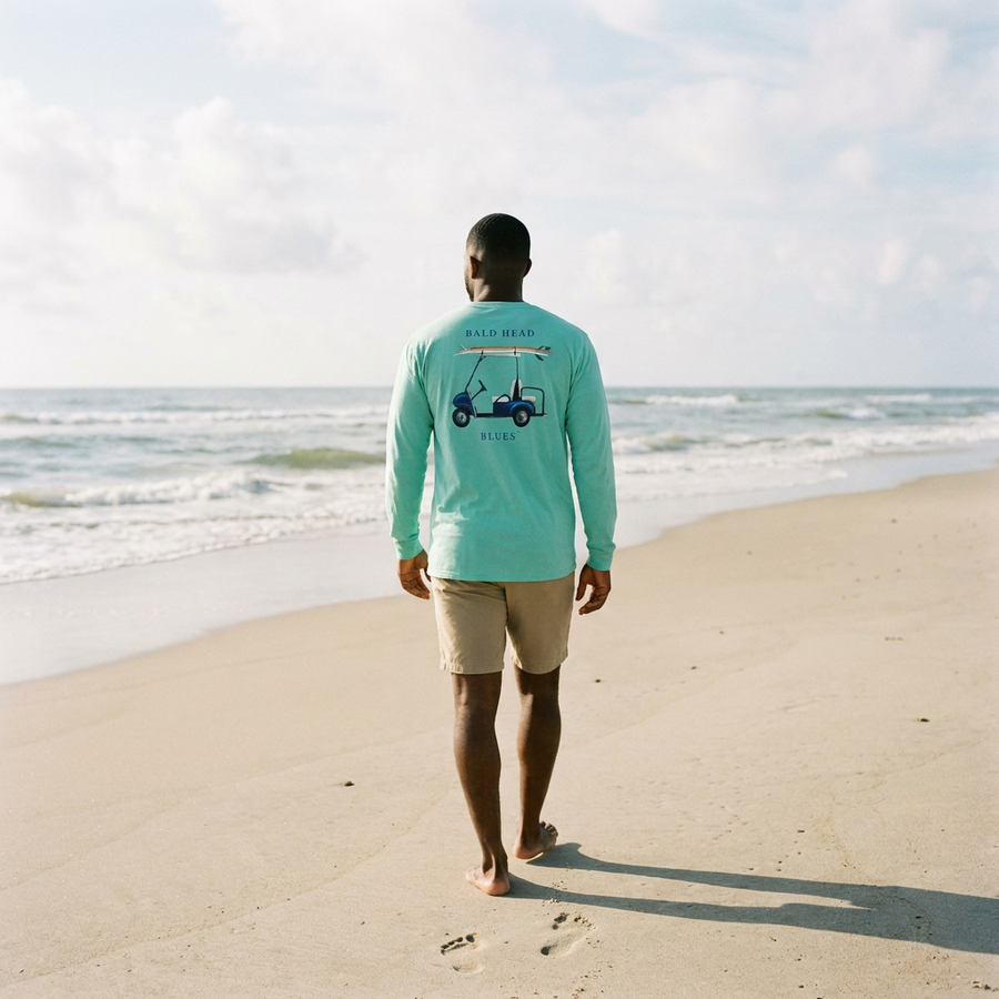 Man in seafoam long-sleeve Island Tee with Bald Head Blues back print walking toward the surf
