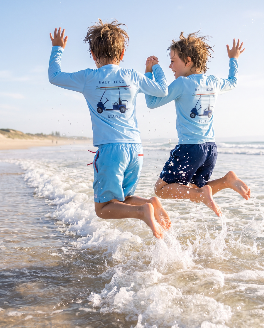 Two boys jumping a wave in Bald Head Blues youth performance golf cart tees and swim trunks